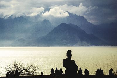 Silhouette people looking at sea and mountains against cloudy sky