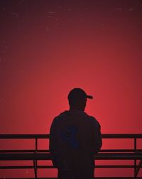 Rear view of man standing by railing against orange sky