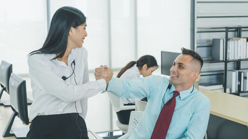 Business people shaking hands with colleague in background at office