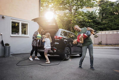 Playful father and son with woman and girl standing by car in back yard