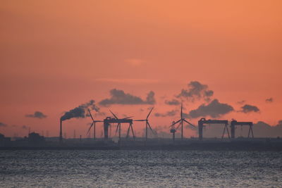 Silhouette factory by sea against orange sky