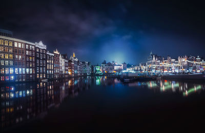 Illuminated buildings by river against sky at night