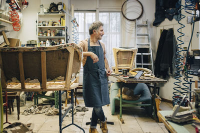 Mature male craftsperson looking away while standing by incomplete sofa in upholstery workshop