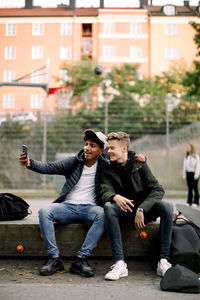 Teenage boy taking selfie with male friend through mobile phone while sitting at park