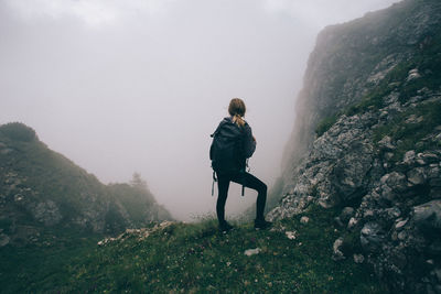 Rear view of man standing on rock against sky during foggy weather