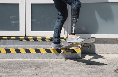 From below young guy with leg prosthesis jumping on skateboard above ground against modern building on sunny day