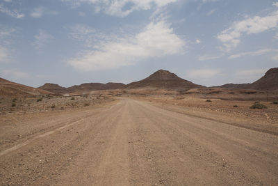 Empty dirt road towards mountains against sky