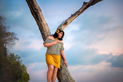 Low angle view of young woman standing by tree against sky