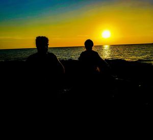 Rear view of silhouette men sitting on beach during sunset