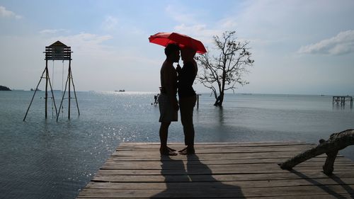 Couple holding umbrella while standing on pier over sea at beach