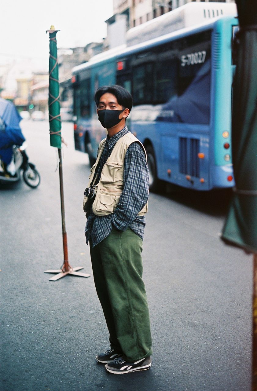 transportation, city, one person, mode of transportation, street, architecture, adult, men, young adult, standing, full length, vehicle, city life, land vehicle, casual clothing, road, travel, blue, day, bus, public transportation, clothing, spring, person, lifestyles, footwear, snapshot, outdoors, portrait, glasses, city street, focus on foreground, train, beard, rail transportation, commuter, leisure activity, fashion, cool attitude