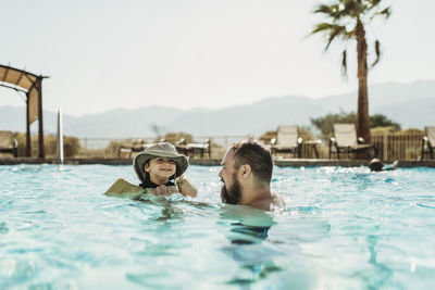 Father and toddler son swimming in pool with mountains and palm trees
