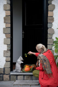 A middle-aged woman in a red cloak with a net of orange pumpkins opens the door