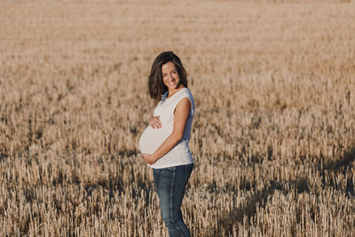 Smiling pregnant woman with hands on stomach standing on field