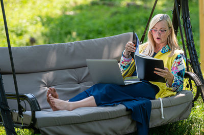 Mature woman with laptop and documents working in garden on rocking couch, green home office concept