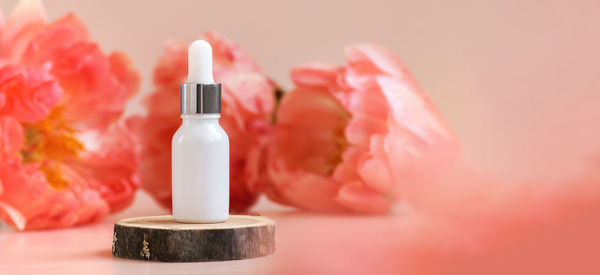 White glass dropper bottle with silver cap on woooden podium with peonies in soft focus