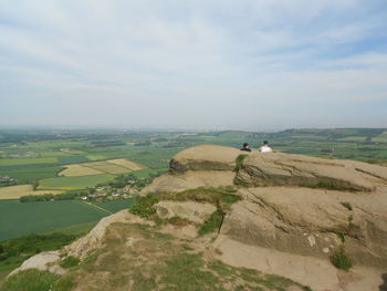 Men sitting on cliff overlooking green landscape against sky