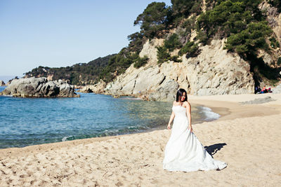 Bride walking at beach against sky during sunny day