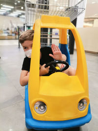 Kid with protective face mask in yellow shopping cart at the mall. 