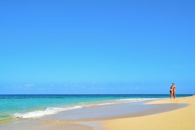 Friends embracing at beach against clear blue sky