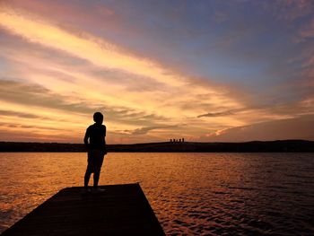 Silhouette boy standing on pier over lake against sky during sunset