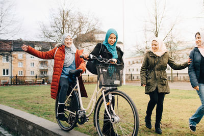 Cheerful multi-ethnic female friends walking with bicycle on grass in city