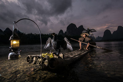 Man with birds sitting on bamboo boat in lake at night