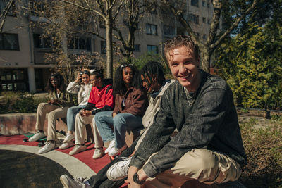 Portrait of smiling young man sitting with multiracial friends on retaining wall in playground