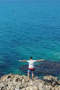 High angle view of man with arms outstretched standing at rocky beach against clear blue sky