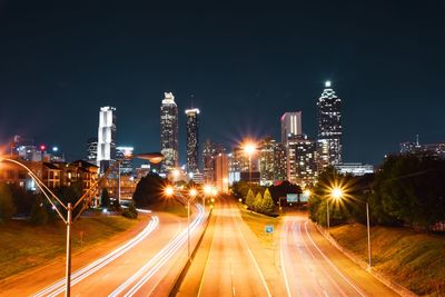 Light trails on road amidst illuminated buildings against sky at night