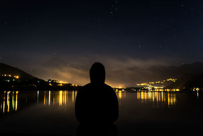 Rear view of silhouette man standing by lake against sky at night