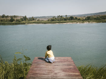 Rear view of boy sitting on pier by lake against sky