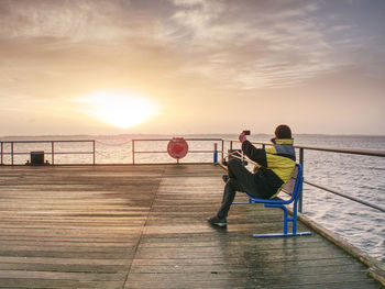 Men sitting on railing by sea against sky during sunset