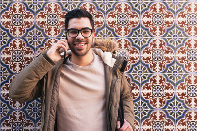 Portrait of man talking on mobile phone while standing against tiled wall