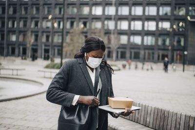 Female entrepreneur with packaged food and laptop standing in office park during covid-19