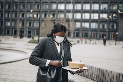 Female entrepreneur with packaged food and laptop standing in office park during covid-19