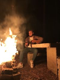 Young man sitting on fire pit at night