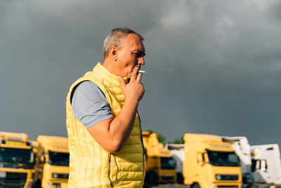 Side view of mature man smoking cigarette against trucks