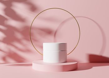 White and blank, unbranded cosmetic cream jar standing on pink podium with golden ring 