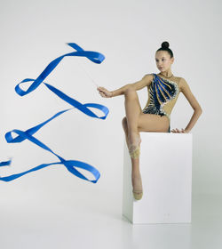 A girl from rhythmic gymnastics in a bodysuit rotates a ribbon while sitting on a cube