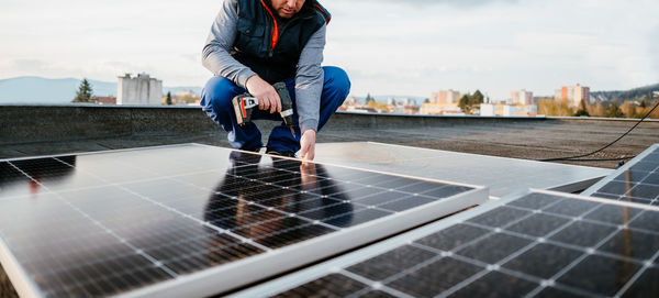 Man fixing solar panel through drill while crouching on rooftop