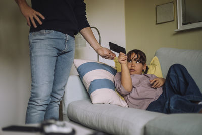 Upset daughter giving smart phone to father standing near sofa at home