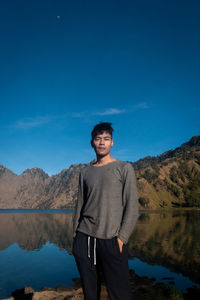 Portrait of young man standing by lake against blue sky