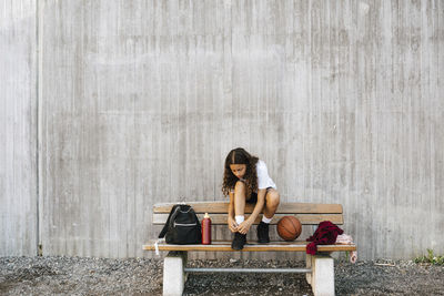 Female basketball player tying shoelace while sitting on bench against wall