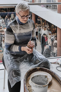 Senior gray-haired woman kneading clay in her hands to sculpt clay vase on potter's wheel.