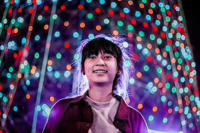 Portrait of smiling young woman against illuminated multi colored lighting equipment