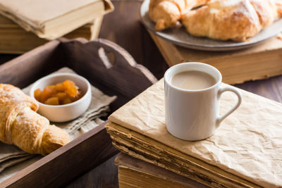 A cup of coffee on a stack of old books and a tray with croissants and jam on a wooden table. 