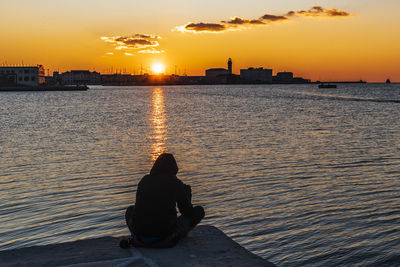 Rear view of woman sitting on shore against orange sky