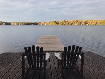 Empty chairs and tables by pier on lake against sky