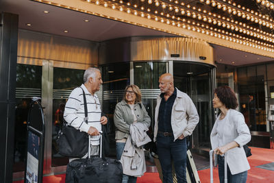 Senior man talking to male and female friends standing with luggage outside movie theater